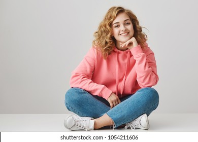 Girl is always happy to hear interesting stories. Curious cheerful woman with curly hair in stylish outfit sitting on floor with crossed legs, leaning on hand, smiling, taking part in conversation