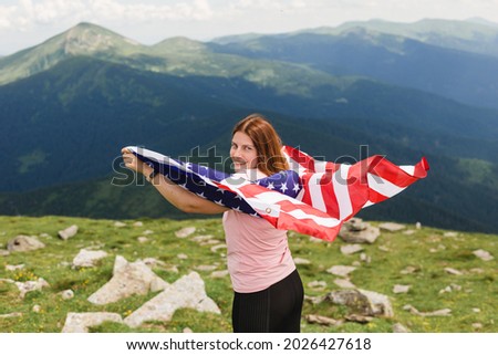 Girl with American Flag looking out at landscape. Young woman stands in the mountains, holding the US flag in her arms high. Flag fluttering in the wind