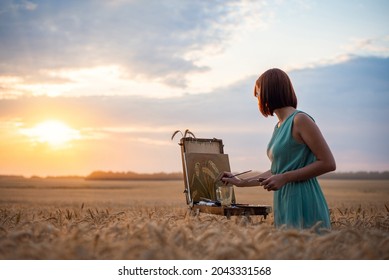 Girl Artist Drawing A Painting In The Field Of Wheat With A Setting Sun In The Background. Beautiful Atmospheric Shot Of A Female Painter Drawing Slill Life Picture With Oil Inks On Canvas