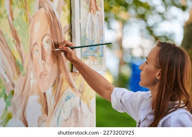 Girl artist draws with paint brush surreal woman portrait on white canvas at outdoor art painting festival, paintings art picture process. Woman in white blouse paints atmospheric surreal picture