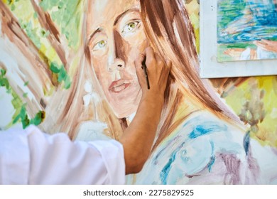 Girl artist hand holds paintbrush   draws surreal woman portrait white canvas at outdoor art painting festival  paintings art picture process  Woman artist paints atmospheric surreal picture