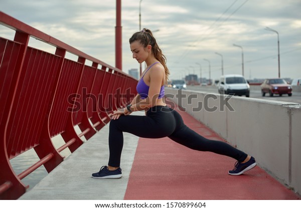 Girl athlete stretching in the\
morning outside, in the city, on modern bridge crossing river.\
Active healthy lifestyle. Sport and recreation\
concept.