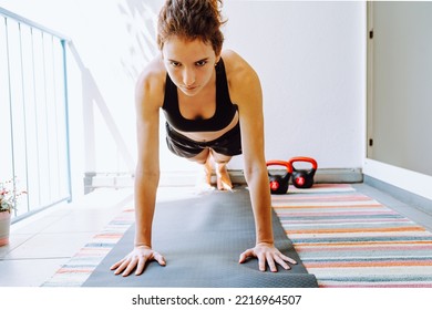 Girl Of Athletic Muscular Build, With Curly Brown Hair Tied In Ponytail, Stands In Plank Position, Pumps Up Arm Muscles, Abs, On Loggia In Sun. Sports Home