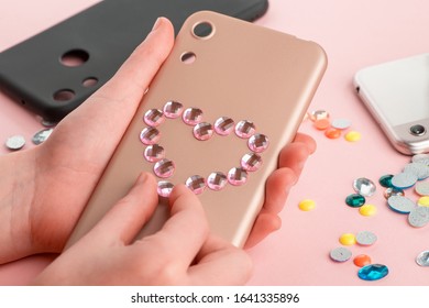 Girl Attaching Pink Rhinestone Heart Onto Beige Phone Case. Romantic DIY Project For Girls. Stylish Phone Case Refashion.