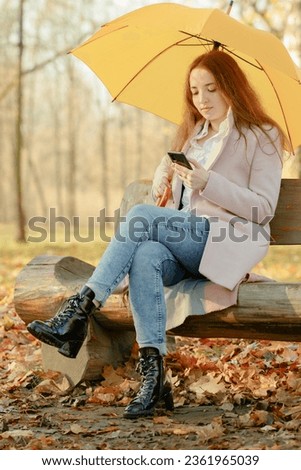A girl in an autumn park sits on a bench carved out of wood, holding an umbrella in one hand and a phone in the other.