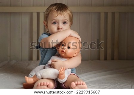 Girl and Baby Doll. Little Girl Playing on a White Blanket in Bedroom. Front View. Child Holding the Doll Toy. Family Relationship Concept. Kid Toddler is Alone, She Hugging and Cradling Her Toy.