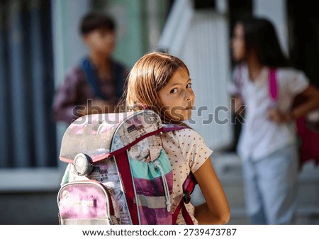 A girl with a backpack looks back while standing in front of a school during the day in front of friends