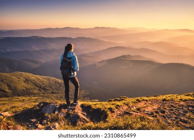 Girl with backpack on mountain peak with green grass looking in beautiful mountain valley in fog at sunset in autumn. Landscape with sporty young woman, foggy hills, orange sky in fall. Hiking. Nature