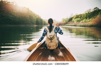 Girl with backpack paddling the wooden canoe on the sunset lake. Rear view of travel woman rowing the boat at sunset