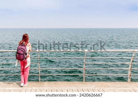 a girl with a backpack stands leaning on a metal fence and looks out to sea. The view from behind. The concept of traveling and active lifestyle. Young woman looking at the sea