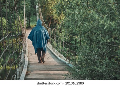 Girl With Backpack Under Blue Raincoat Is Engaged In Hiking. On Summer Evening At Sunset, She Walks Along Wooden Suspension Bridge Over River. Concept Of Savage Travel, Lifestyle, Outdoor Recreation