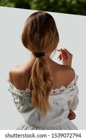 A Girl With Bare Shoulders In A White Top Is Standing With Her Back. Long Hair Is Collected By A Ribbon With Pearls. The Photo Is Taking On A White Background With A Green Stripe At The Top.