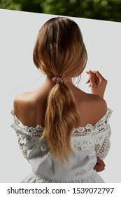 A Girl With Bare Shoulders In A White Top Is Standing With Her Back. Long Hair Is Collected By A Ribbon With Pearls. The Photo Is Taking On A White Background With A Green Stripe At The Top