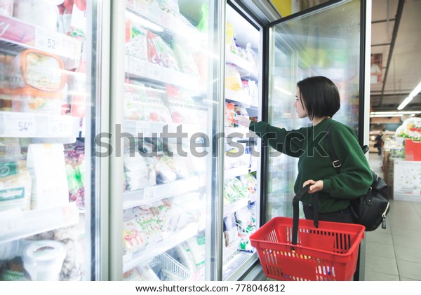 A girl with a basket takes frozen foods from\
the supermarket\'s refrigerator. The girl chooses goods in the\
store. Shopping in the\
supermarket.