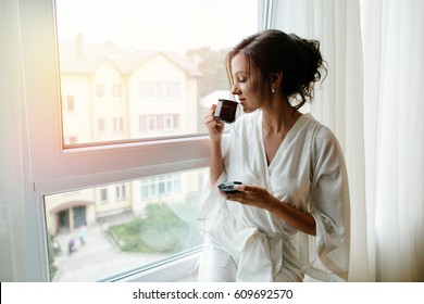 A girl in a bathrobe drinking coffee in the morning on the windowsill.