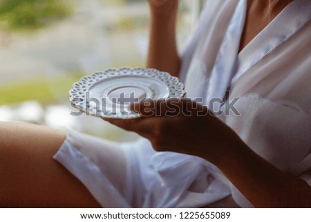 Girl in a Bathrobe sits on the windowsill next to the window and drinks coffee from a mug with a saucer. Young woman in a bathrobe drinking hot espresso or cappuccino in the morning on the windowsill.