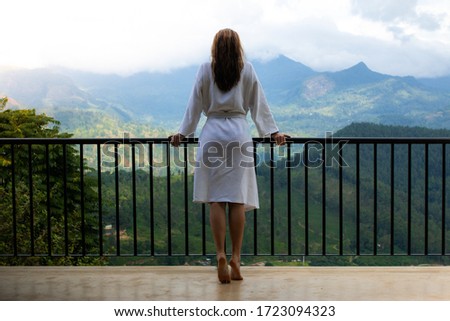 Girl in bathrobe standing at a balcony looking / watching over a mountain range during sunrise