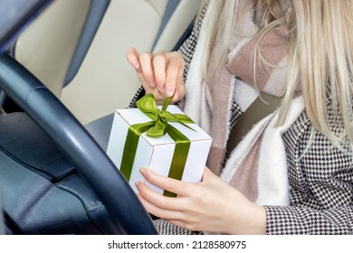 Girl With Beautiful Blonde Hair Color Is Sitting In A Car, On The Driver Seat And Is Unpacking A White Gift Box, Green Ribbon. Girl Next To Steering Wheel, Pastel Clothes Colors,cute Girl. Smile