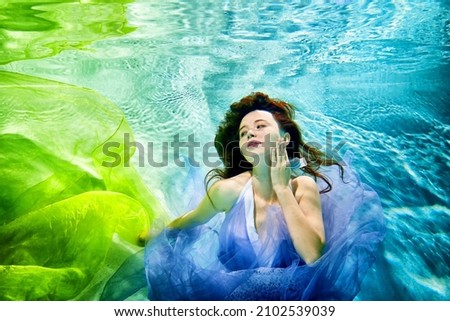 Girl in a beautiful dress under blue water. Female model posing under water in blue swimming pool