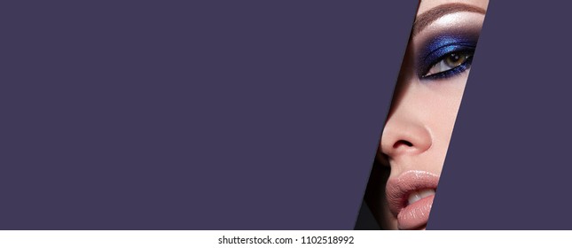 A girl with beautiful green bright eyes with blue shining shadows, beige lipstick and expressive eyebrows looks into the hole of colored paper.Fashion, beauty, make-up, cosmetics, beauty salon, style.