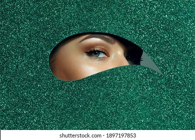 A Girl With Beautiful Green Eyes, With Bright Shadows On Her Eyelids. Looks Through A Teardrop Hole Made Of Green Shiny Paper.