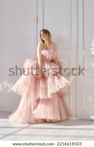 A girl in a beautiful lush long dress of the color of a dusty rose stands near a light wall. Vintage bridal glamor. Fashion shooting in a wedding or evening dress