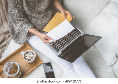 Girl in beige knit sweater and white pants sitting at home in cozy atmosphere and is holding golden envelope.In lap of girl is laptop. Girl using gadget. In the background white knitted pillow.