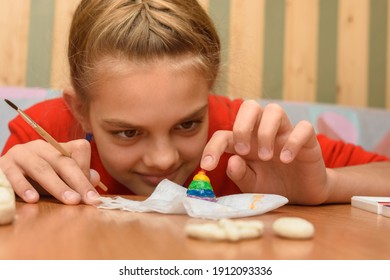 A Girl Bending Over Looks At A Painted Figurine Made Of Salt Dough