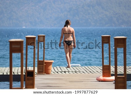 Girl in bikini going to swim in sea on wooden pier on mountain background. Beach vacation, summer leisure by the sea
