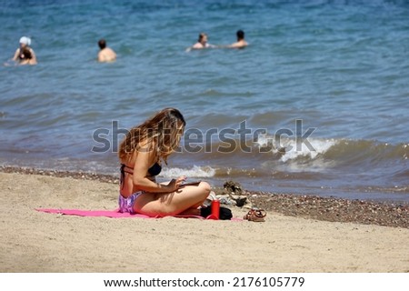 Girl in bikini sitting and sunbathing on sea beach with smartphone in hand on swimming people background. Summer vacation, online communication on resort