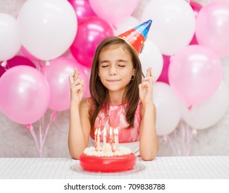 Girl in the birthday hat with a cake with candles makes a wish with his fingers crossed
