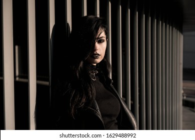 Girl in black with black aggressive make up with is standing behind white striped wall in a city. Might be a picture representing subculture, youth, teenagers, evil, protest