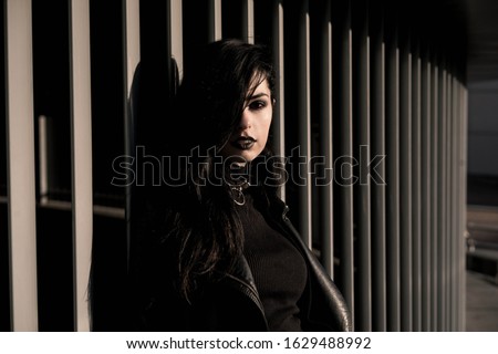 Girl in black with black aggressive make up with is standing behind white striped wall in a city. Might be a picture representing subculture, youth, teenagers, evil, protest