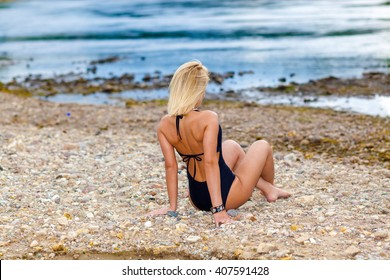 Girl In A Black Bikini Swimsuit On The Stony Beach. Sexy Blonde Woman Sunbathing. View From The Back 