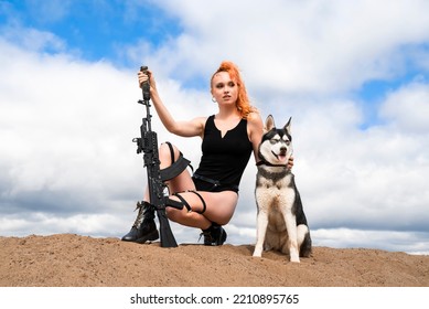 A Girl In A Black Bodysuit With Bare Legs Holds An Assault Rifle In Her Hands While Sitting On The Sand. A Woman With A Siberian Husky Dog On The Background Of A Blue Cloudy Sky.