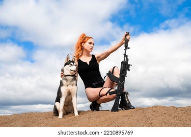 A Girl In A Black Bodysuit With Bare Legs Holds An Assault Rifle In Her Hands While Sitting On The Sand. A Woman With A Siberian Husky Dog On The Background Of A Blue Cloudy Sky.