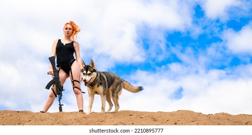 A Girl In A Black Bodysuit With Bare Legs Holds An Assault Rifle In Her Hands While Standing On The Sand. A Woman With A Siberian Husky Dog On The Background Of A Blue Cloudy Sky.