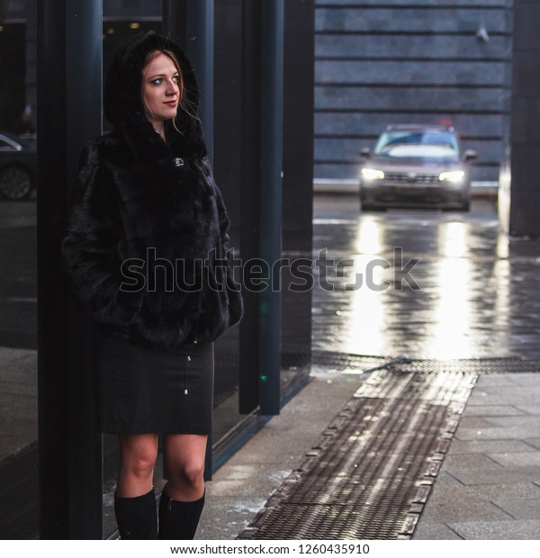 A girl in a black coat in cold\
weather in the business center of the city in the light of the\
headlights of a parked car. Emotion of sadness and\
reflection.