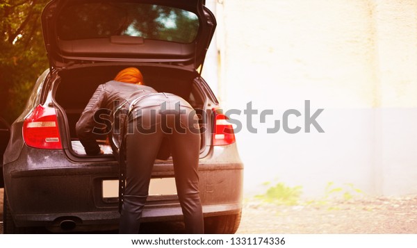 Girl in a black leather pants and suit,\
back. The girl rummaging in the trunk of the\
car