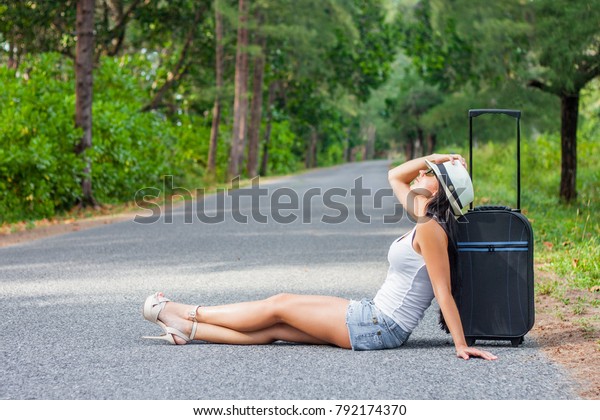 a girl black
long hair in hat glasses shorts and heels, with a suitcase is get
tired and sitting  alone with hope on the road on the roadside on
hot sun and waiting car 