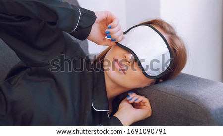 Girl in black pajamas on the gray sofa. Young woman sleeps with a blindfold on her eyes. Phone and flowerpot on the table. String bag on a white wall.