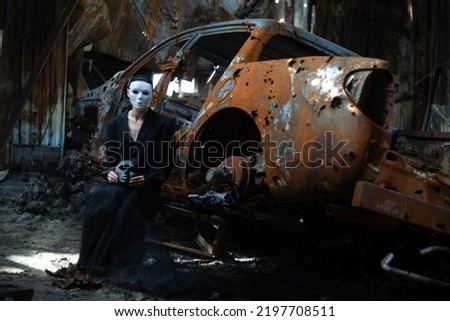 A girl in a black suit and a white mask sits near a burnt-out car from a Russian army shell hitting the house.