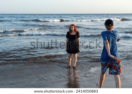 A girl in a black summer dress stands in the waves by the sea, a boy in shorts and a cap runs to her