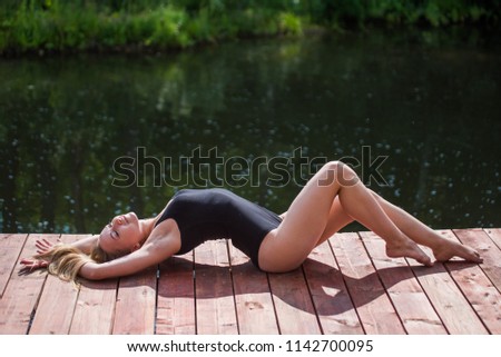 Girl in a black swimsuit lying on her back by the lake. Elegant slender young blond woman in black swimwear enjoying a relaxing day at the lake lying on her back on a wooden jetty in the sunshine