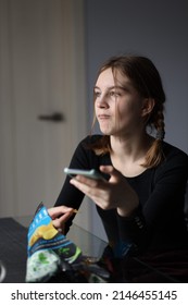 Girl In Black T-shirt Sits In Kitchen Holding Phone In Her Hand And Eating Chips