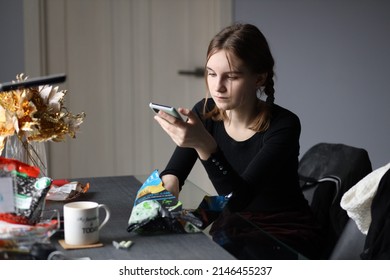 Girl In Black T-shirt Sits In Kitchen Holding Phone In Her Hand And Eating Chips