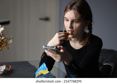 Girl In Black T-shirt Sits In Kitchen Holding Phone In Her Hand And Eating Chips