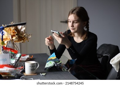 Girl In Black T-shirt Sits In Kitchen Holding Phone In Her Hand And Eating Chips