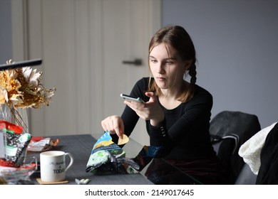 Girl In Black T-shirt Sits In Kitchen Holding Phone In Her Hand And Eating Chips