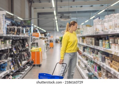 A Girl With Blond Hair Of European Appearance In Jeans And A Yellow Jacket Stands Near The Shelves In A Store With A Blue Plastic Cart